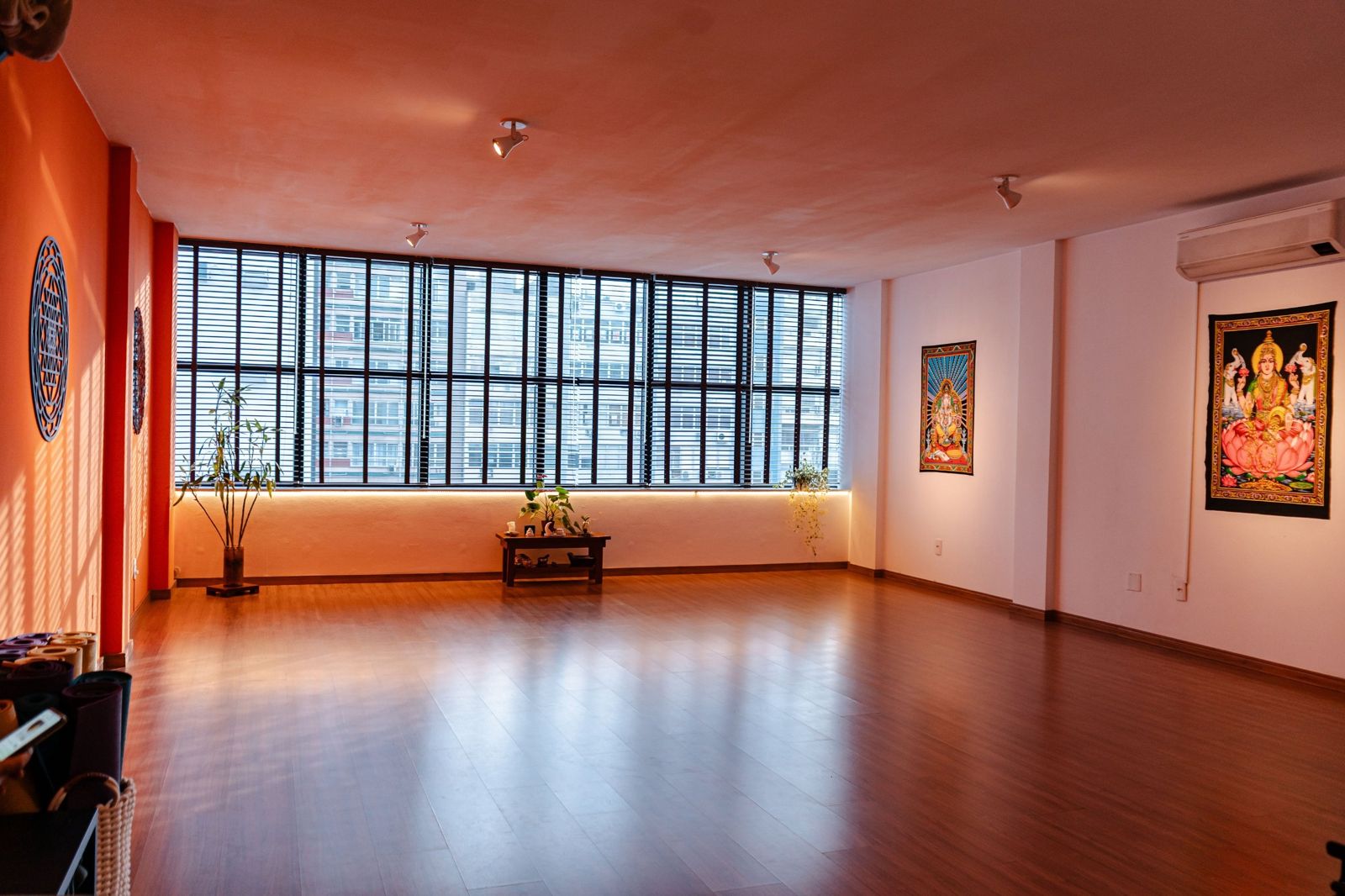 Sala da Surya Yoga vazia e ampla, com a parede laranja, as janelas e a parede branca com quadros